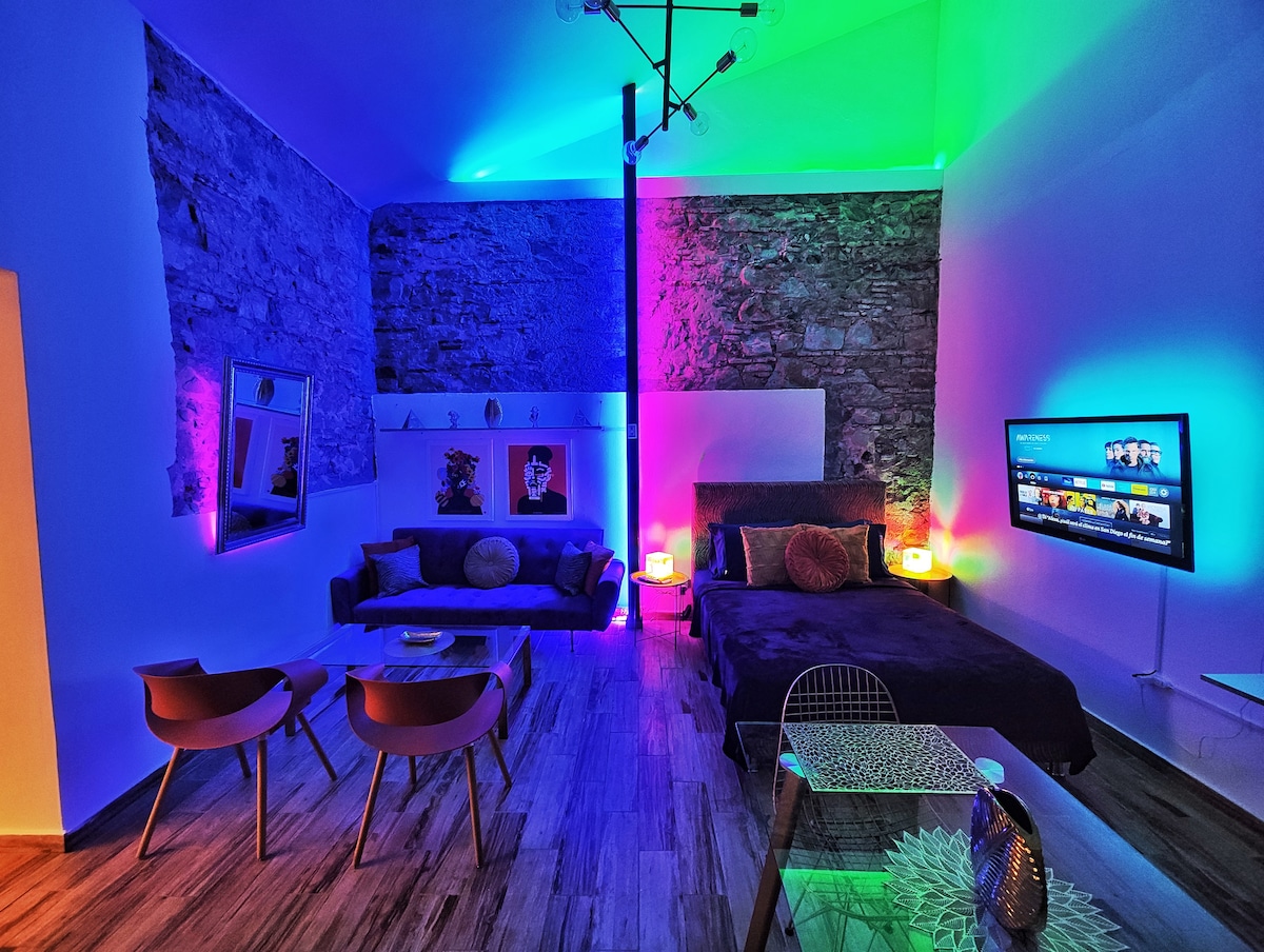 A modern living space showcases exposed stone walls and stylish lighting in vibrant colors. A spacious bed complements a cozy seating area with two chairs. A wall-mounted television adds convenience, while a glass table enhances the open feel of the room.