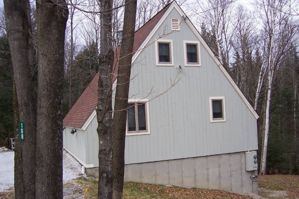 A cozy chalet is showcased with a sloped roof and light grey siding. Large windows are visible on the upper level, blended with surrounding trees. The structure sits on a slight incline, with a gravel path leading to the entrance and a natural, wooded backdrop.