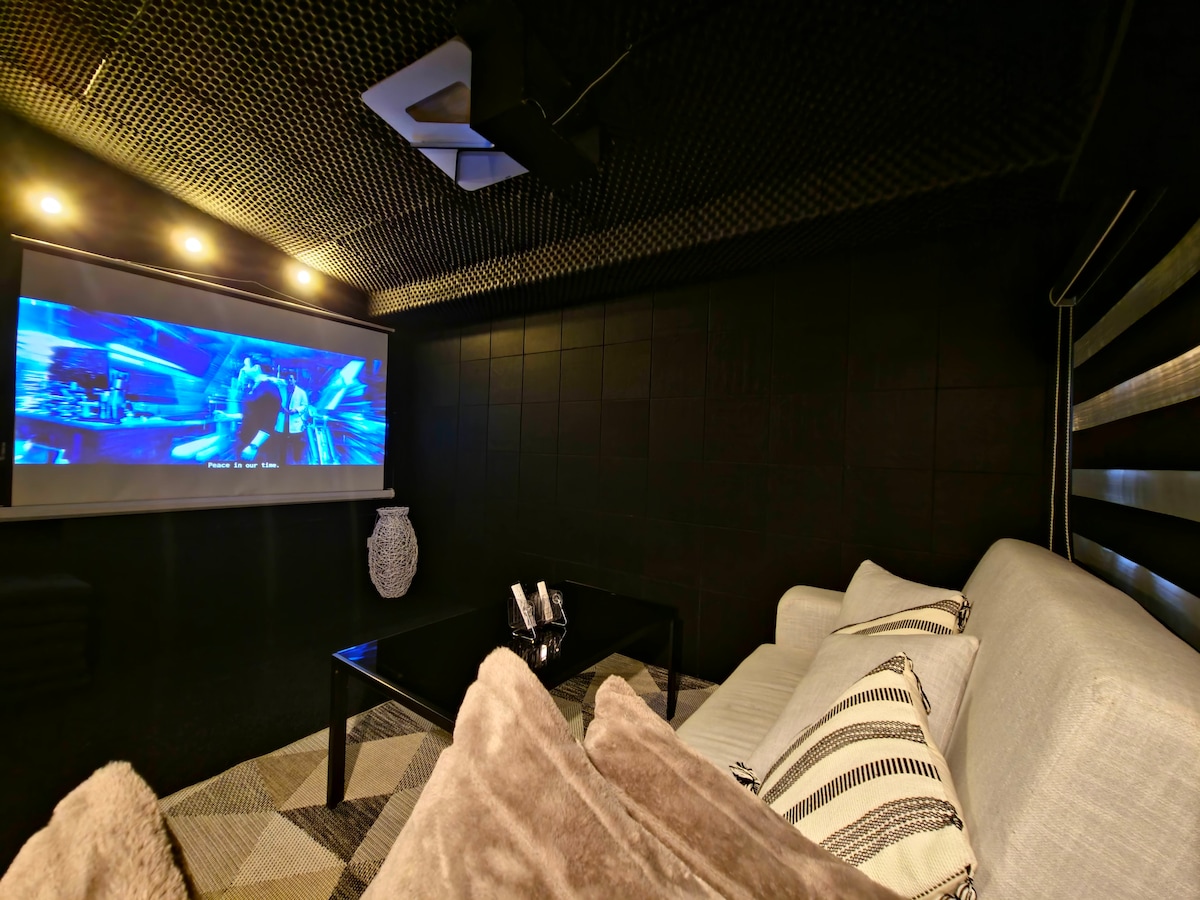 A cozy theater/game room is equipped with a large projector screen displaying a movie. A comfortable sofa with plush cushions is positioned in front, complemented by a sleek coffee table. Soft lighting enhances the inviting atmosphere, suitable for entertainment or relaxation.