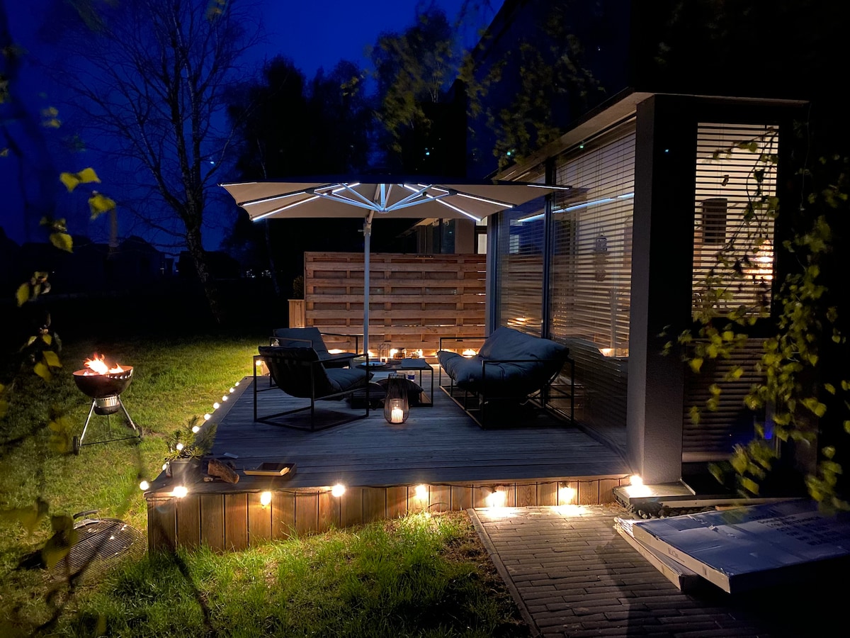 An outdoor terrace is softly illuminated by surrounding lights, providing a calming evening ambiance. Comfortable lounge furniture is arranged under a large umbrella, with a small fire pit nearby. The grassy area offers a serene environment, enhanced by the natural surroundings.
