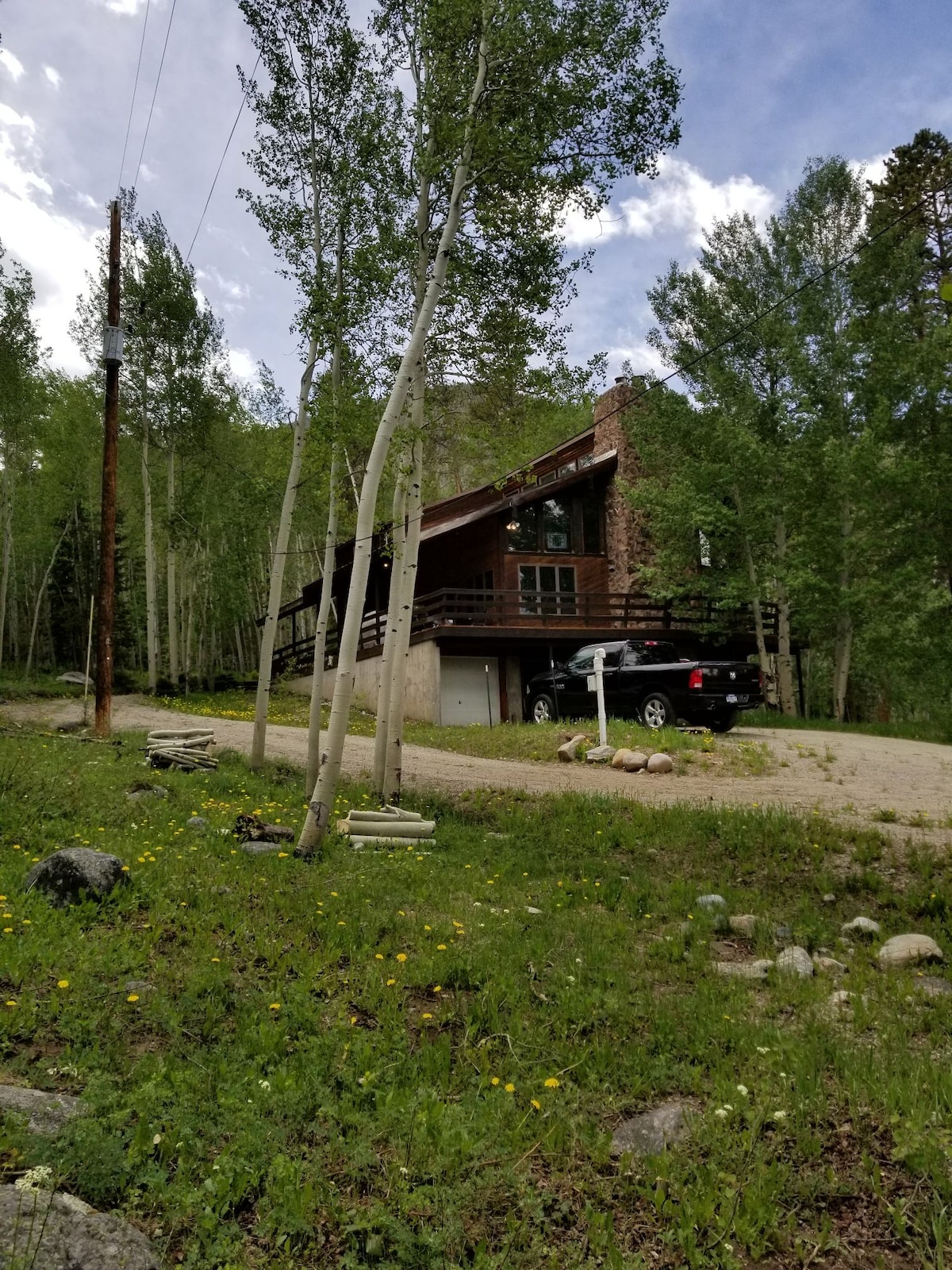 A large rustic cabin is set among lush greenery, featuring a two-story structure with a sloping roof. A gravel driveway leads to the front entrance, and a dark vehicle is parked nearby. Tall trees surround the property, creating a secluded ambiance.