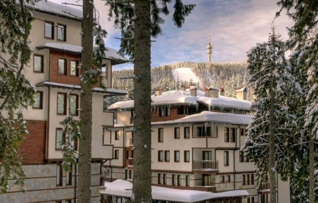 A multi-story building features a combination of wood and stone elements under a blanket of snow. Surrounding pine trees are visible, adding to the alpine setting. In the background, a tower can be seen on the snow-covered hillside.
