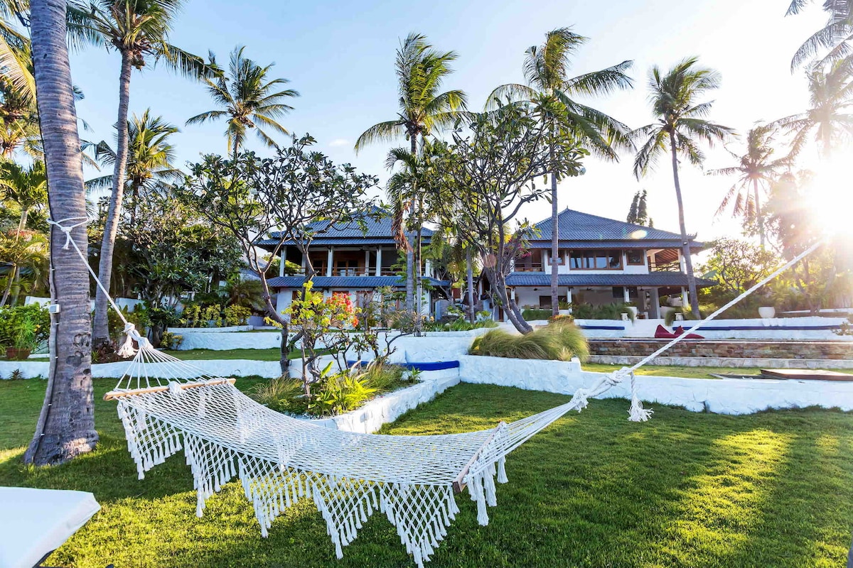 A white hammock is centrally positioned on well-manicured green lawn, surrounded by tropical palm trees and vibrant plants. The villa's structure is visible in the background, framed by sunlight that enhances the serene outdoor setting.