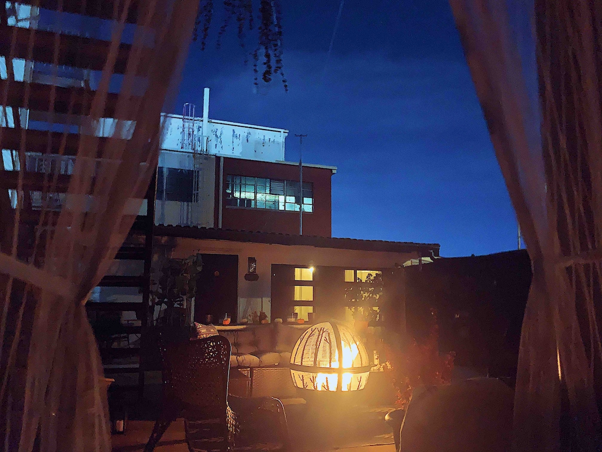 An outdoor area is illuminated by a soft glow from a fire pit, surrounded by comfortable seating. The silhouette of a building stands against a deep blue evening sky, framed by sheer curtains that gently sway in the breeze.