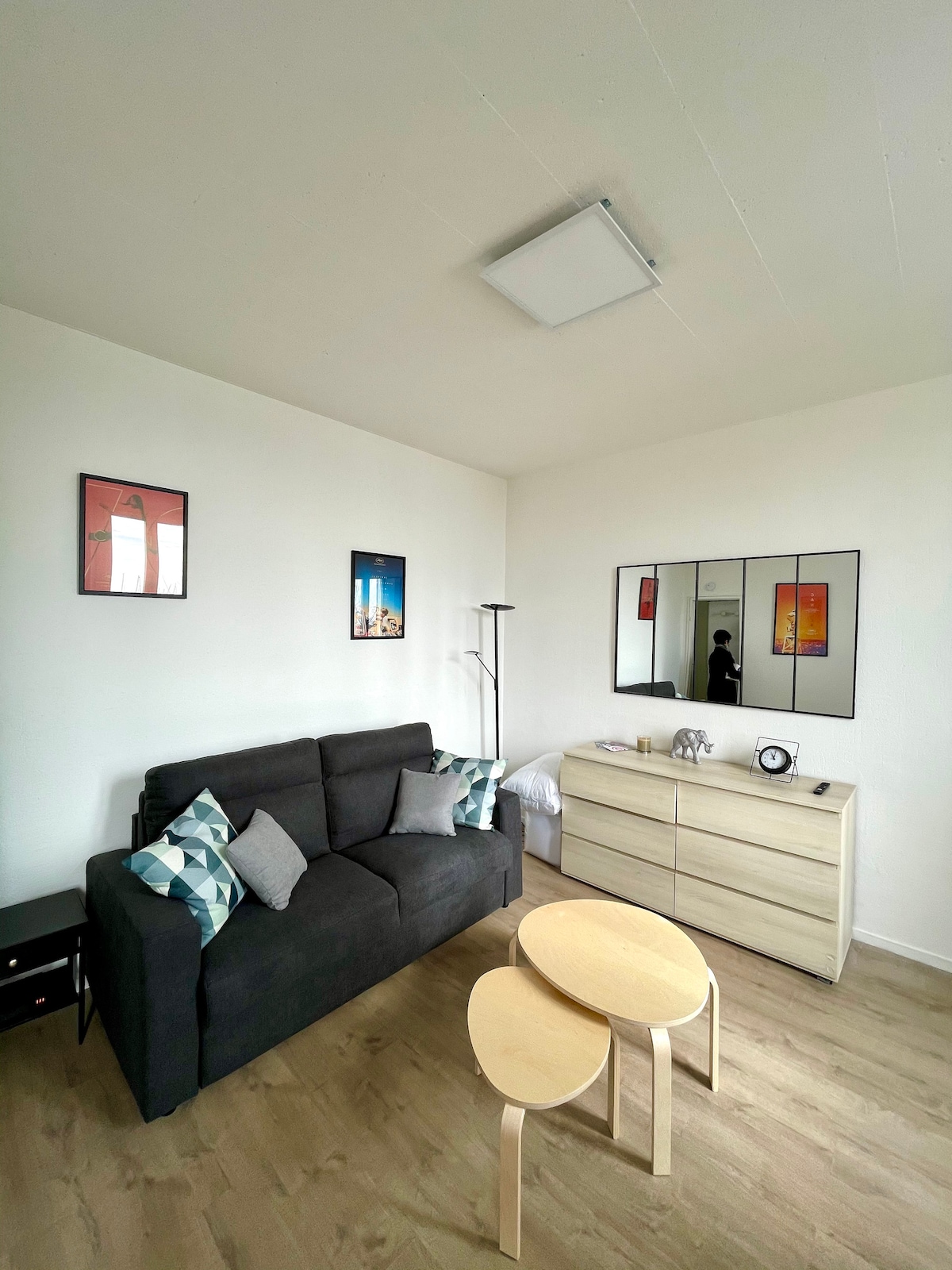 A comfortable lounge area features a dark sofa adorned with patterned cushions. Two light wooden tables are placed in front of the sofa. A large, framed mirror reflects the simplicity of the space, while two colorful artworks are mounted on the walls, adding character.