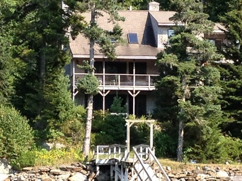 A coastal home is set among tall trees, featuring a spacious deck that overlooks a deep water dock. The structure showcases a mix of wood and stone, with large windows reflecting natural light. A pathway leads to the dock, welcoming access to the water.
