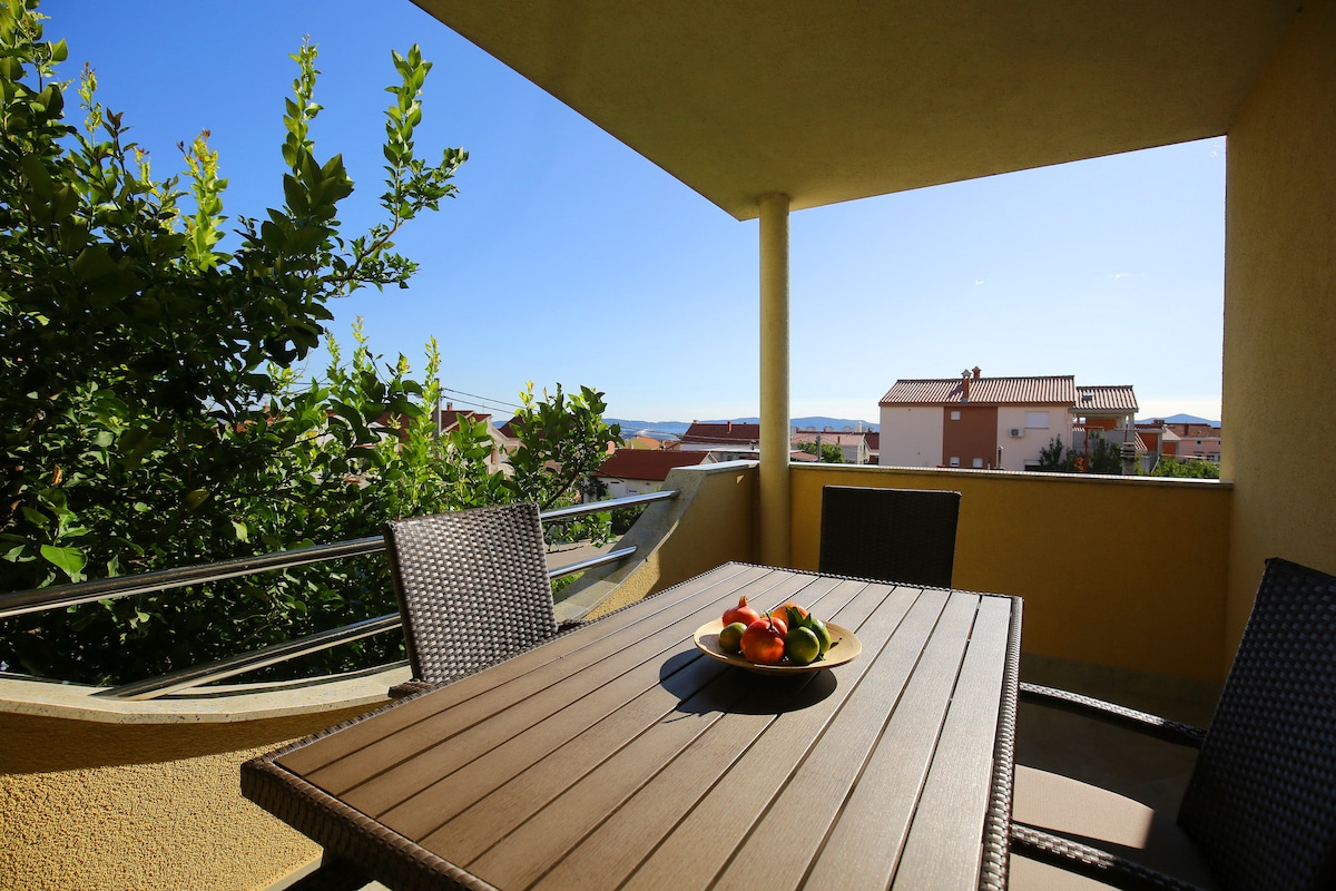 A terrace is featured with a wooden dining table set for four, accompanied by stylish woven chairs. A fruit bowl sits at the center of the table. Lush greenery surrounds the space, offering views of the neighborhood and distant landscapes under a clear blue sky.