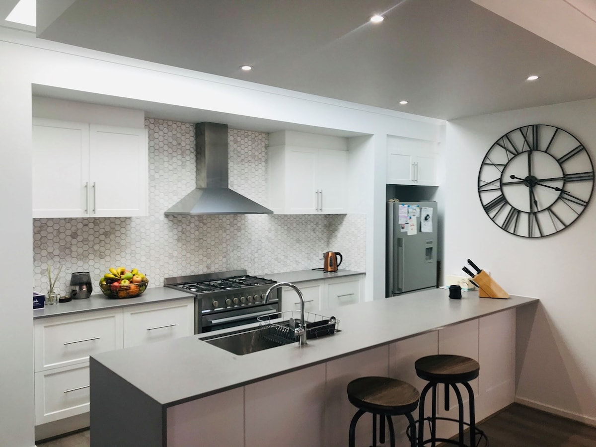 A modern kitchen features sleek white cabinetry and a geometric tiled backsplash. A stainless steel range hood sits above a gas stove, with a large countertop that includes a sink. Bar stools are positioned alongside the island, and a contemporary clock adorns the wall.