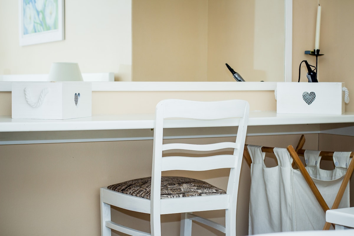 A simple white desk is depicted with a matching chair featuring a patterned seat. Two storage boxes are placed on the tabletop, and a candle holder is visible. A mirror reflects the soft, neutral-colored walls, while a clothes rack is positioned in the background.