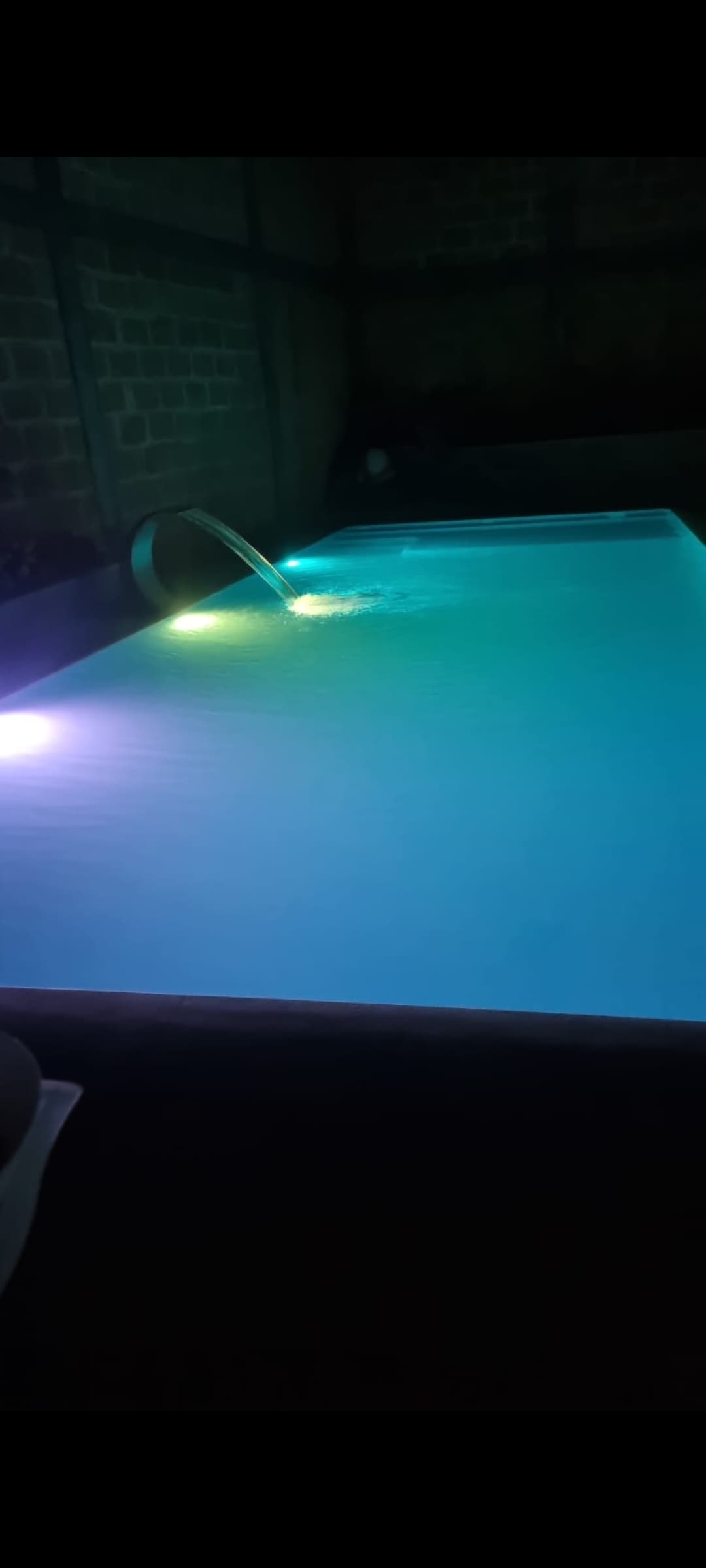 A tranquil pool is illuminated with soft, colored lights creating a calming atmosphere. The water surface reflects shades of blue and green, enhancing the serene setting. The surrounding area is dimly lit, promoting a peaceful ambiance for relaxation.