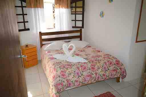 A well-lit sleeping area is furnished with a bed adorned in floral bedding. Towel swans are arranged decoratively on the bed. A window draped with light curtains allows natural light to fill the room, while a small bedside table is positioned next to the bed.