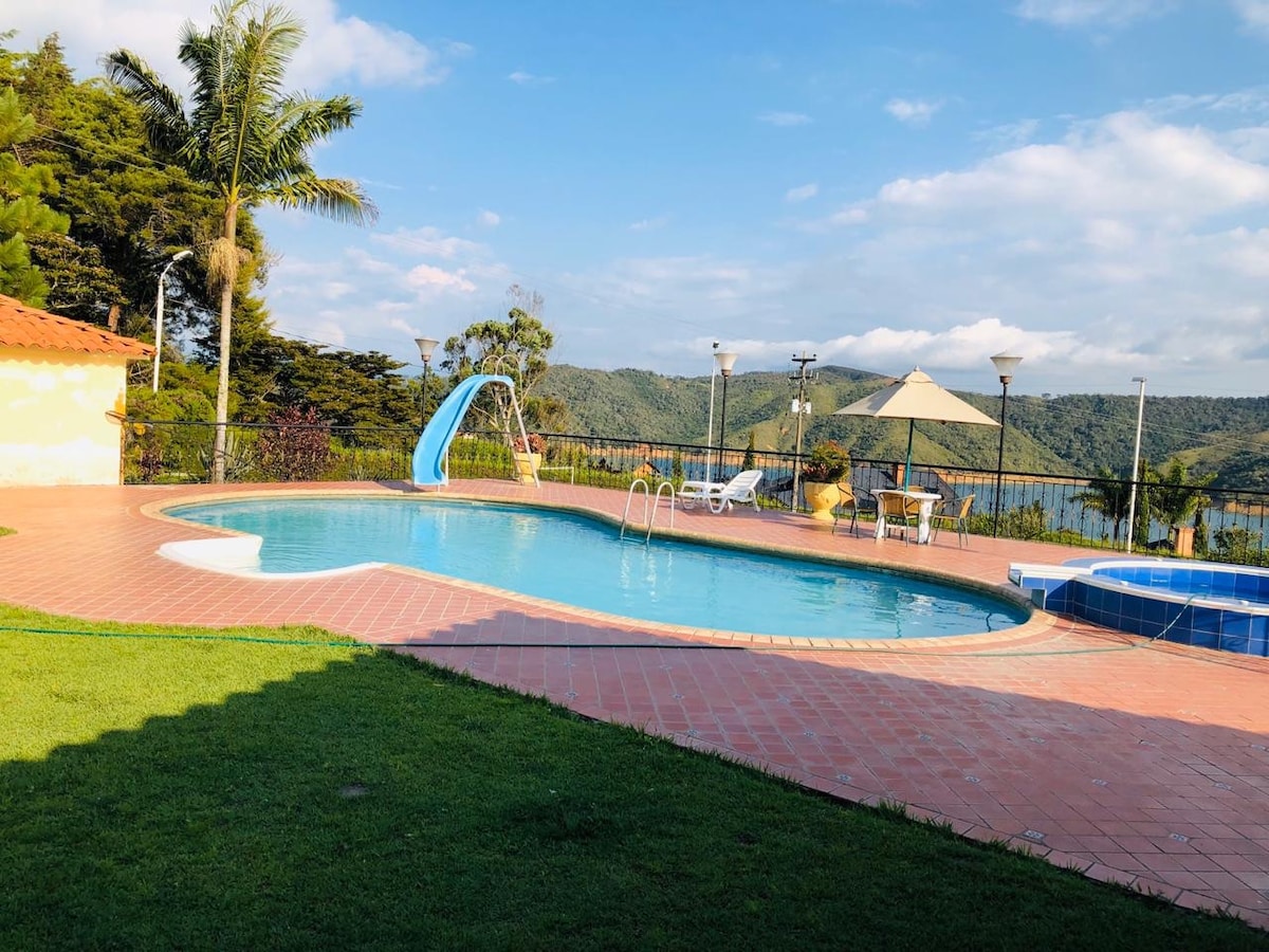 A pool area features a curved swimming pool with a water slide and a hot tub nearby. Sun loungers and round tables are positioned under a shaded gazebo. Lush green grass surrounds the space, and a scenic view of hills and a partly cloudy sky is visible in the background.