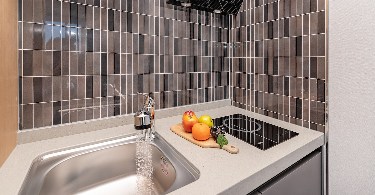 A modern kitchen area is highlighted by a sleek countertop and a stainless steel sink. A cutting board displaying assorted fruits rests nearby, while a two-burner cooktop is integrated into the surface. The backsplash features a mosaic of dark tiles, contributing to a contemporary design.