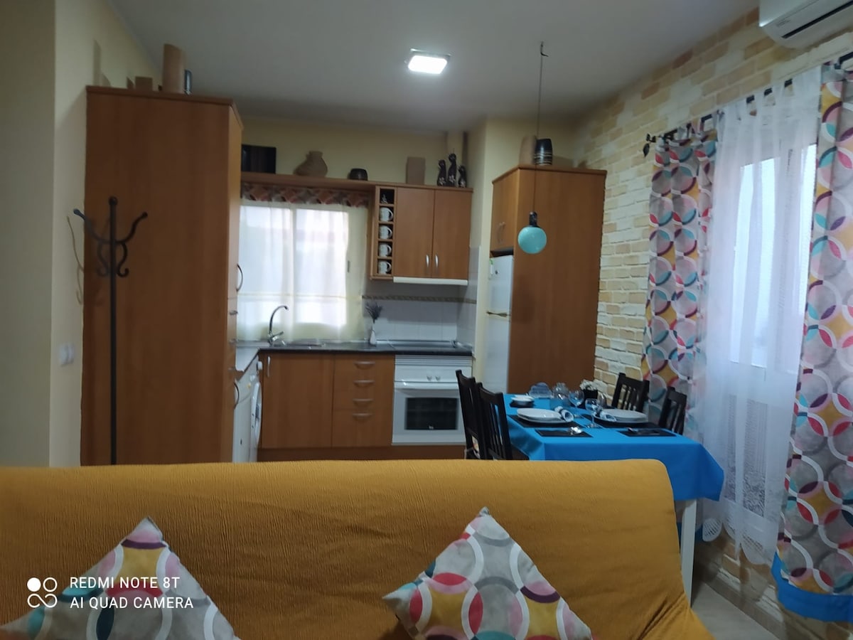 A well-appointed kitchen and dining area are visible, featuring wooden cabinetry and a kitchen island. A dining table set for four is draped with a bright blue tablecloth, and natural light filters through sheer curtains, illuminating the space.