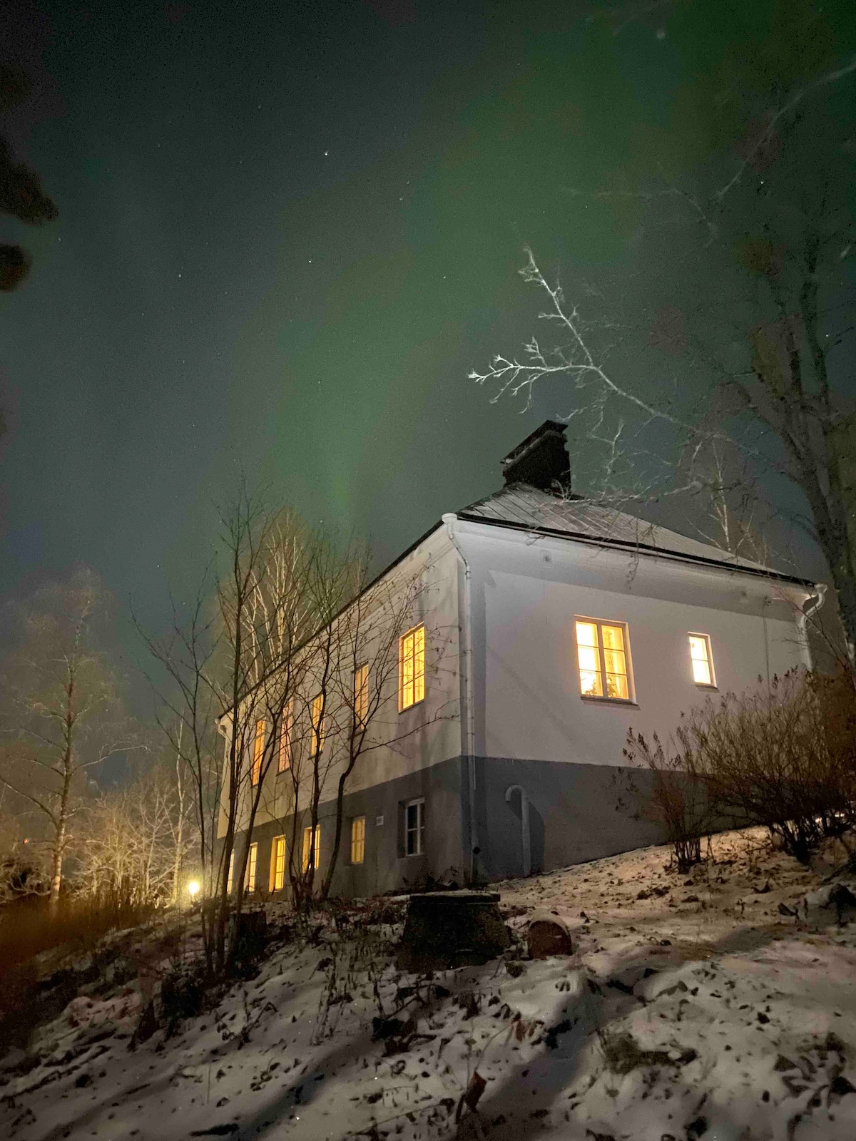 The exterior of the villa is illuminated by warm light from several windows, contrasting against the snowy ground and surrounding trees. The clear night sky features the northern lights, creating a calming ambiance over the quiet landscape.