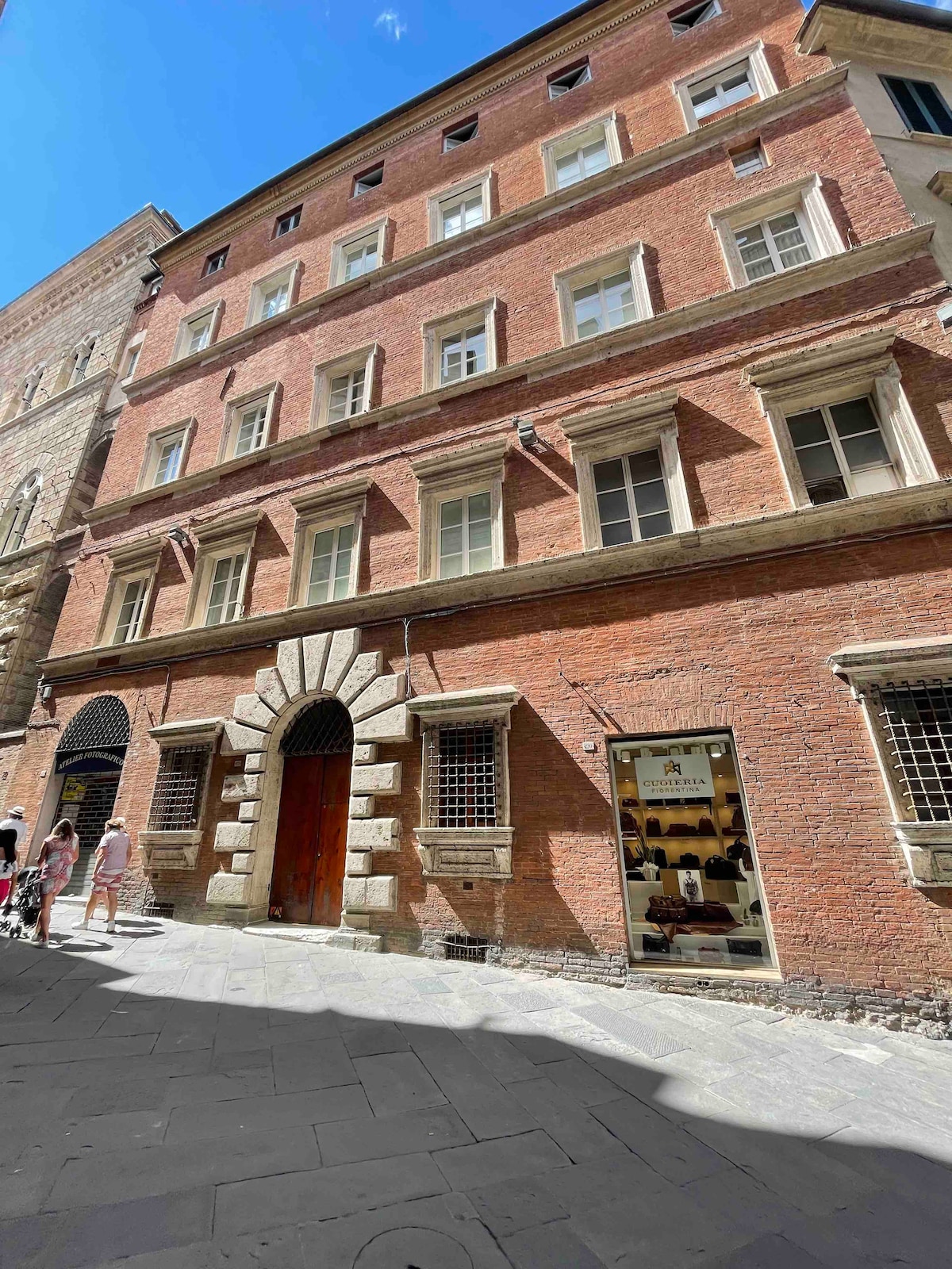 The exterior of a historic brick building features a decorative arch above a wooden door. Windows are positioned symmetrically along the upper levels, complemented by stone accents. The lively street scene includes pedestrians, with adjacent structures showcasing traditional architecture and blue skies above.