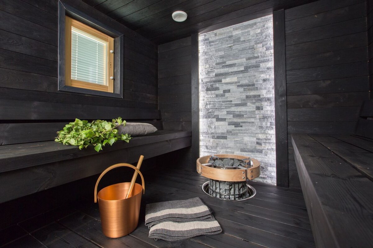 The sauna features dark wooden walls, creating a calming environment. A stone wall with a modern design is visible next to a fire pit surrounded by stones. A wooden bucket and a folded towel are placed nearby, contributing to the authentic sauna experience.
