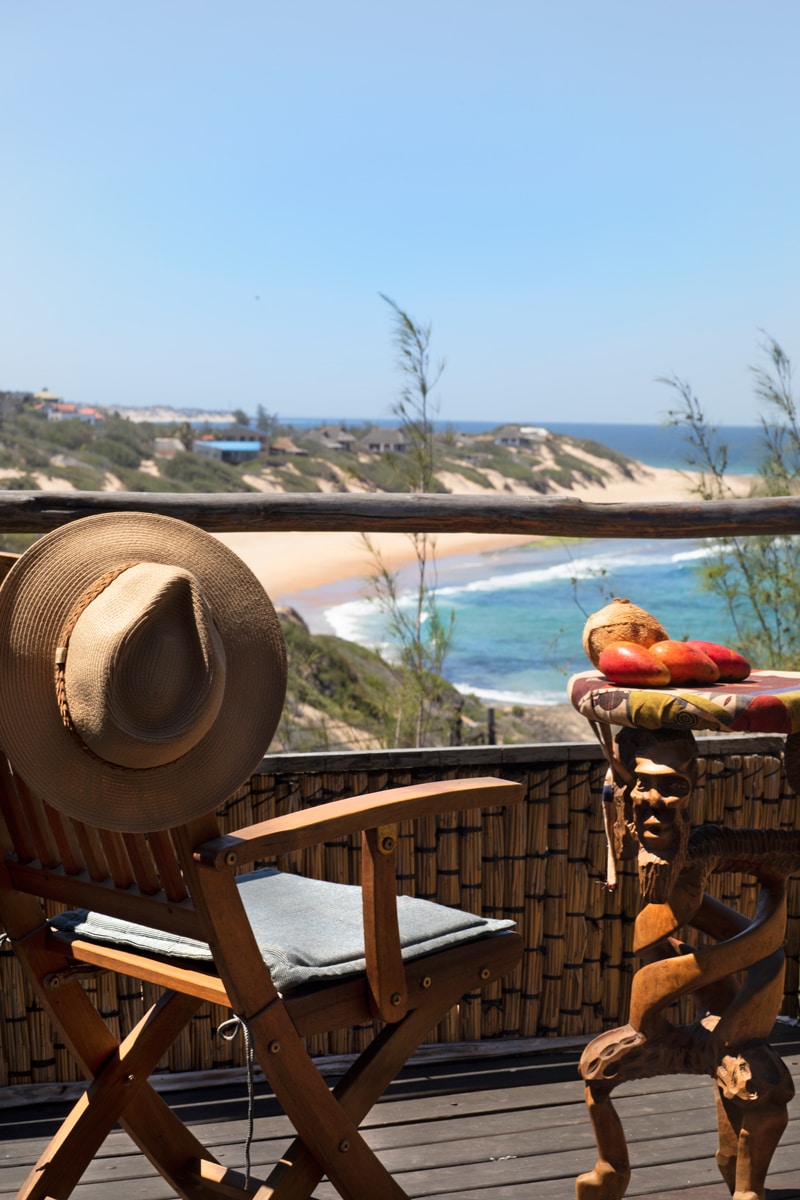 A wooden chair adorned with a sun hat overlooks a scenic view of Tofinho beach. The coastline curves gently, with soft sand meeting the vibrant blue of the ocean. A small table, featuring local craftsmanship, holds a few ripe fruits, adding a touch of color to the serene setting.