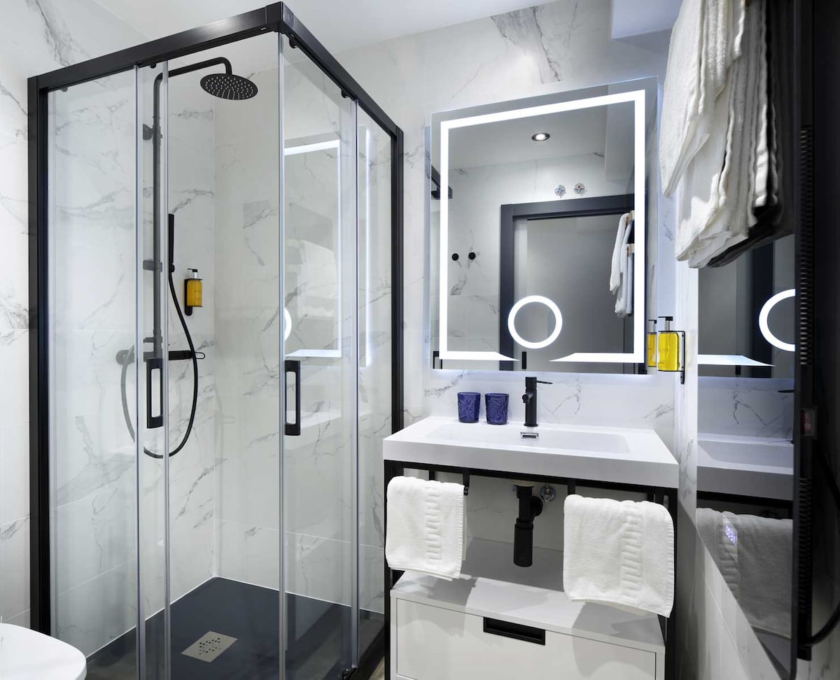 An elegant bathroom features a glass-enclosed shower with modern fixtures. A well-lit mirror is framed in LED, reflecting a stylish vanity with dark accents. White towels and toiletries are neatly arranged, contributing to a clean and organized space.