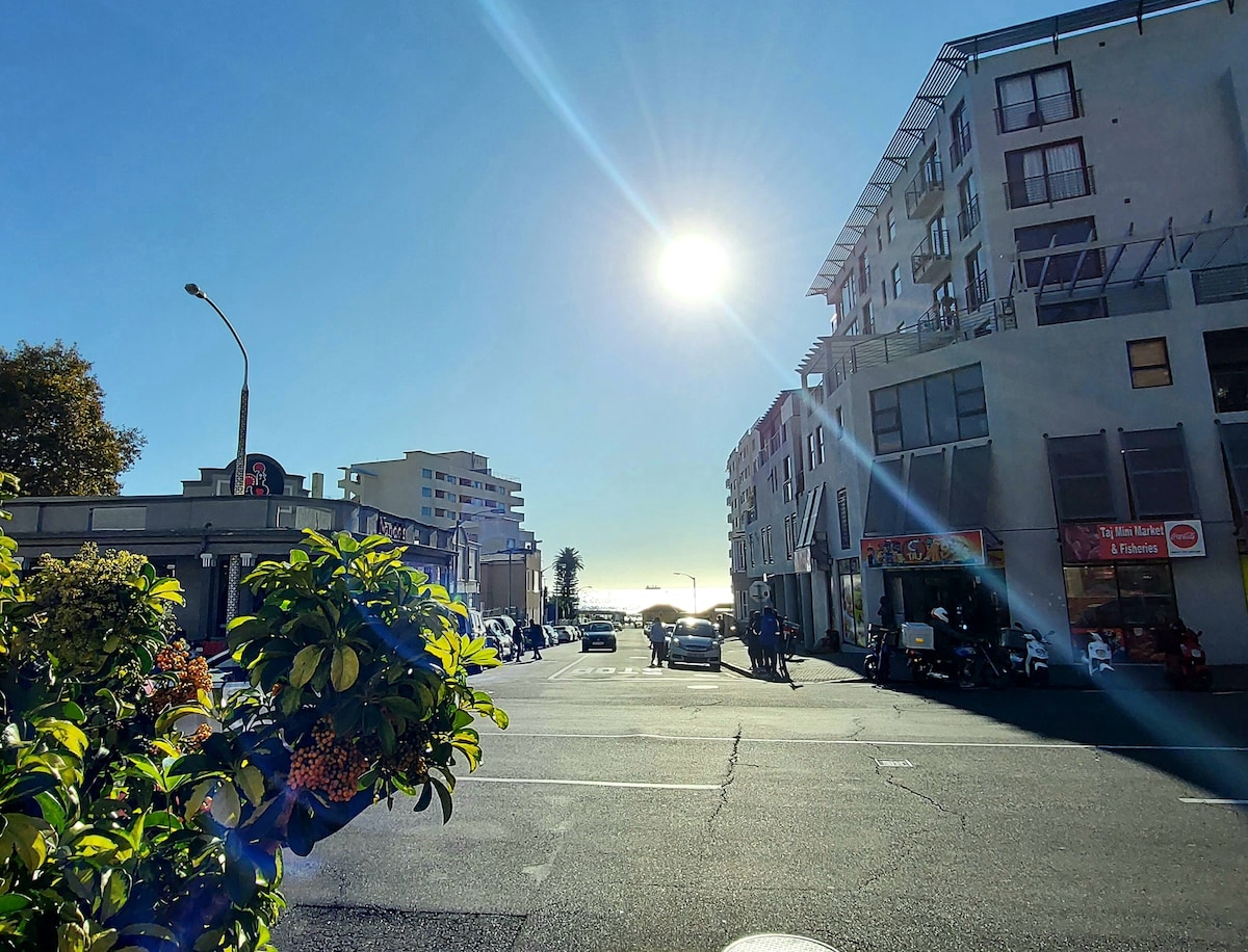A clear street view captures a sunlit day in Sea Point. Buildings line the road, showcasing a mix of architectural styles. Lush greenery in the foreground adds a vibrant touch, with the Atlantic Ocean visible in the distance. The scene conveys a lively urban atmosphere.