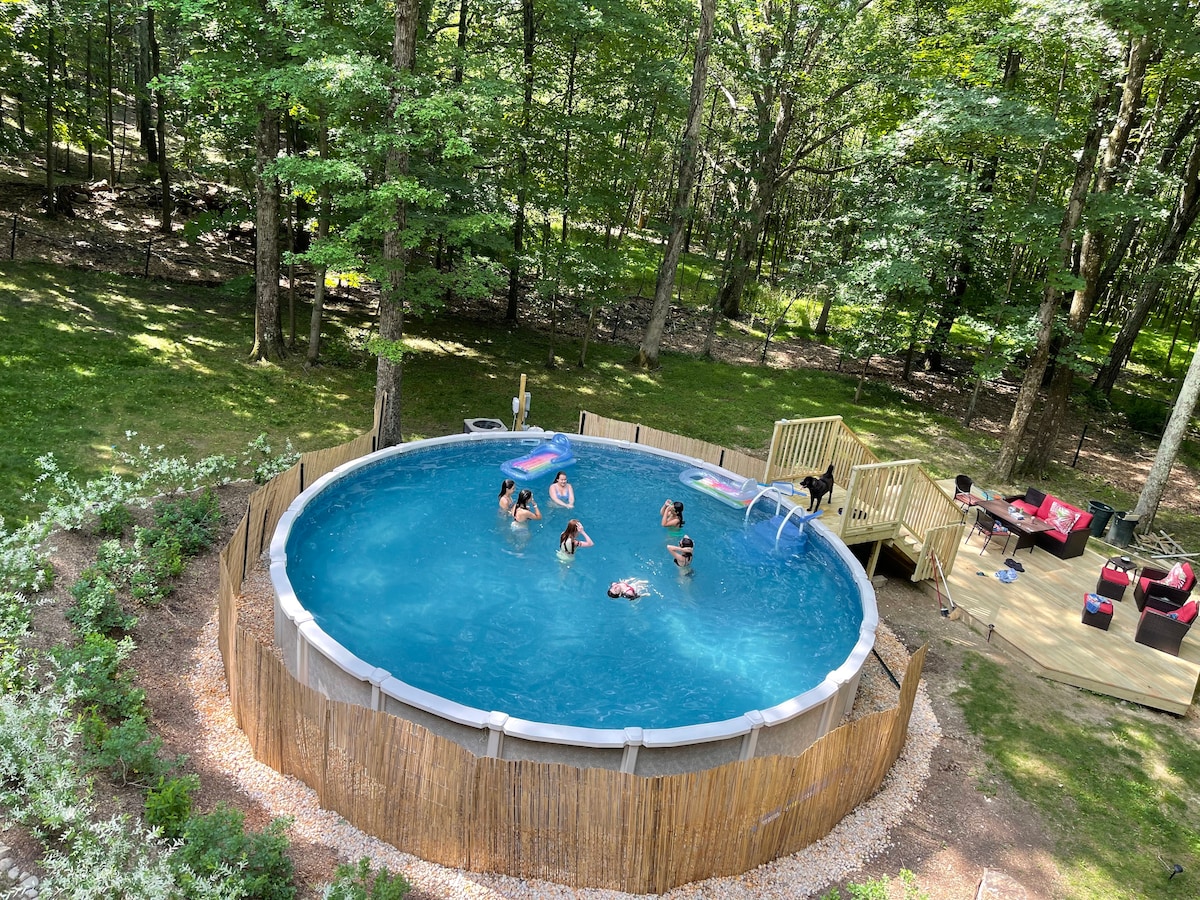 A heated saltwater pool, surrounded by a wooden deck, is set within a fenced yard surrounded by trees. Colorful pool floats and lounge chairs are visible, while guests enjoy the refreshing water in a serene forested environment.