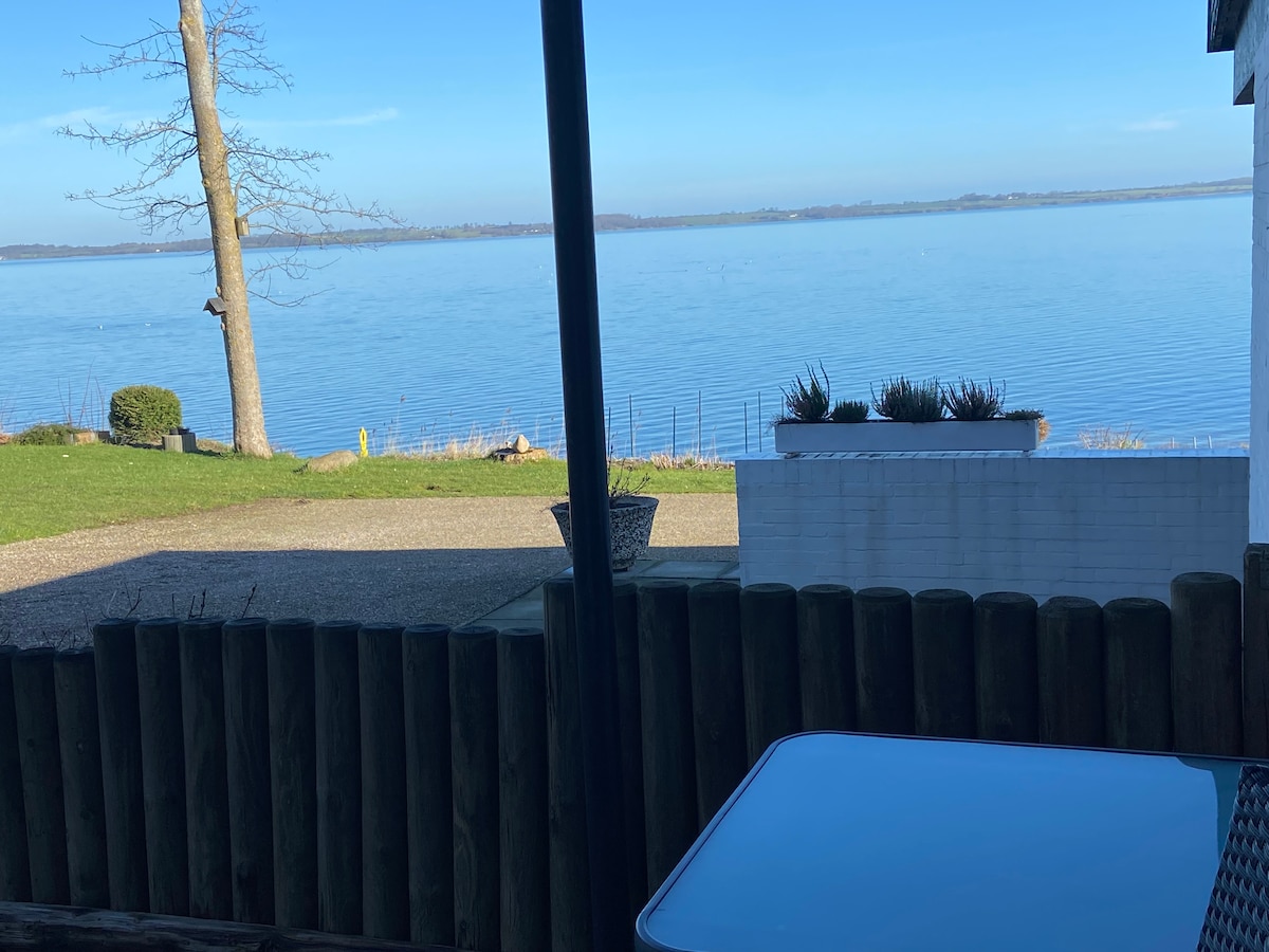 A view from the terrace is captured, showcasing calm waters reflecting the sky. Green grass and a small garden area are visible, along with a wooden fence and a table. The distant shoreline can be seen along the horizon, contributing to a serene atmosphere.