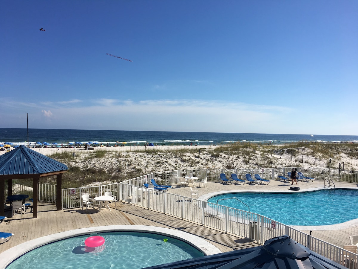 A clear view of the beach and ocean can be seen beyond two pools surrounded by lounge chairs. A covered gazebo is positioned nearby, adding to the outdoor space. The beach features soft white sand and umbrellas, inviting relaxation under the sun.