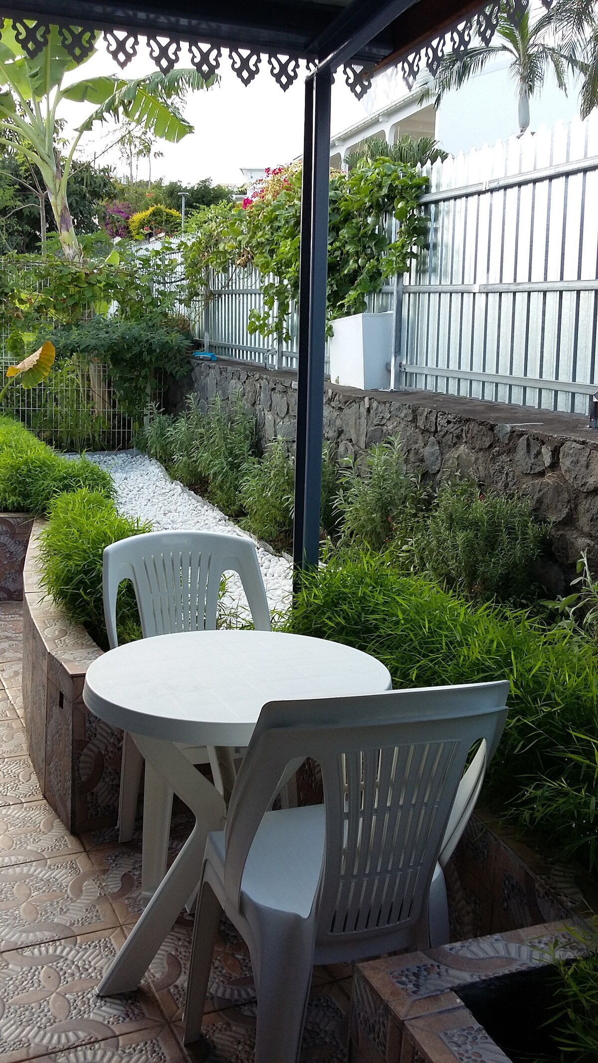 A small outdoor seating area features a round table paired with two chairs under a shaded canopy. Lush greenery surrounds the space, complemented by a neatly arranged pathway of white stones, creating a serene environment for relaxation.