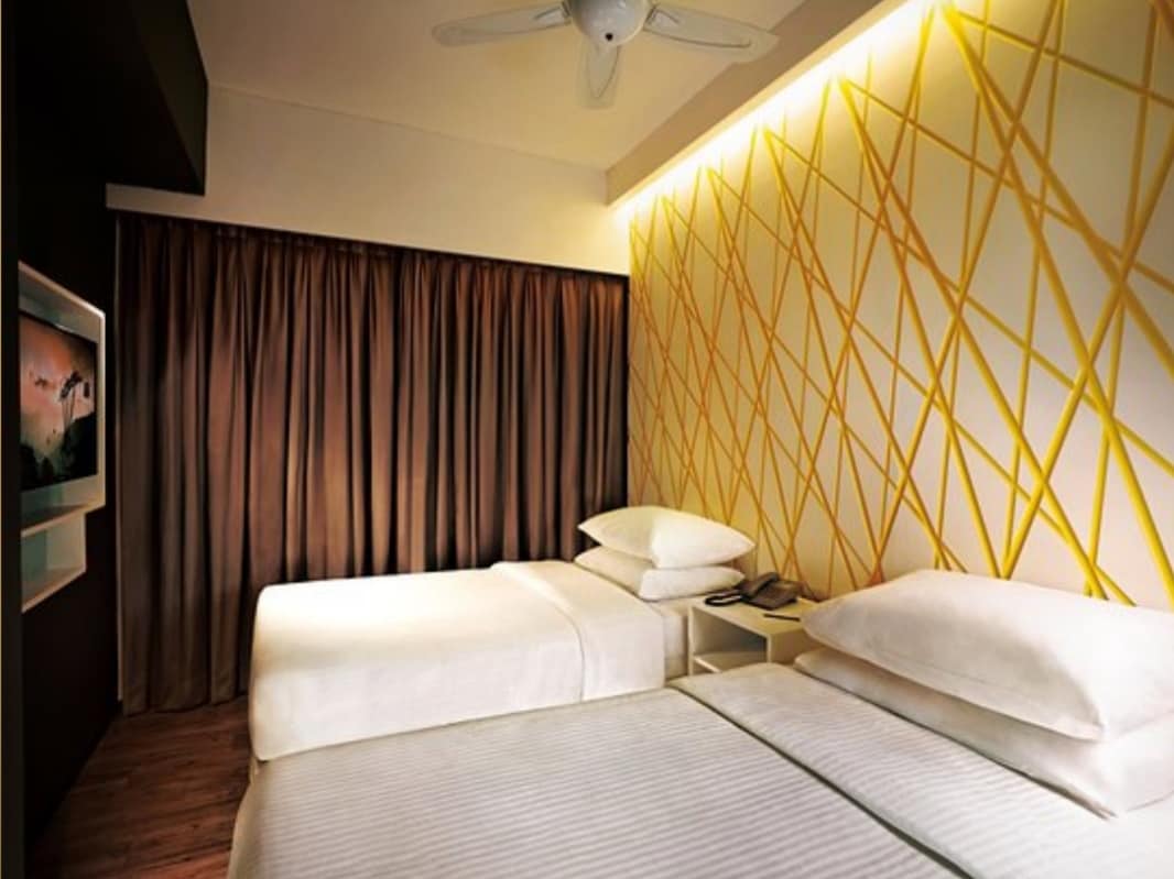 A well-appointed room displays two spacious beds with crisp white linens. A patterned wall features vibrant yellow lines, and soft brown curtains frame a window. A ceiling fan enhances airflow, while a mounted television is positioned for easy viewing.