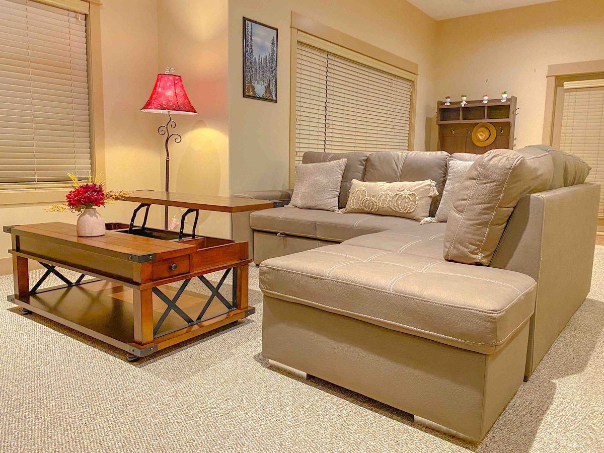 The living room features a modern sectional sofa in neutral tones, accompanied by a wooden coffee table with a lift-top for dual function. A decorative lamp provides soft lighting, while large windows allow natural light to fill the space.
