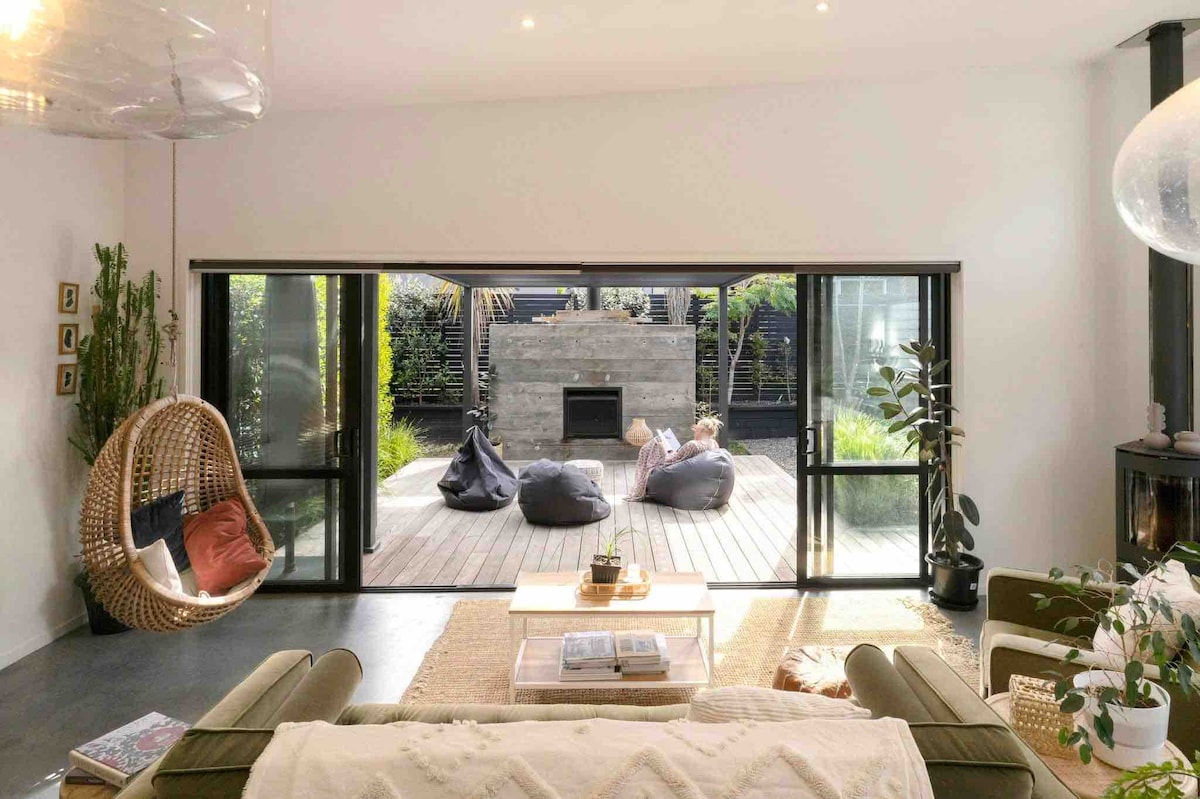 A spacious living area features large glass doors opening to a private deck, creating an airy connection to the outdoors. Cozy seating includes a swinging chair and comfortable couches, with soft textiles adding warmth. A fireplace is set against a stone wall in the background.