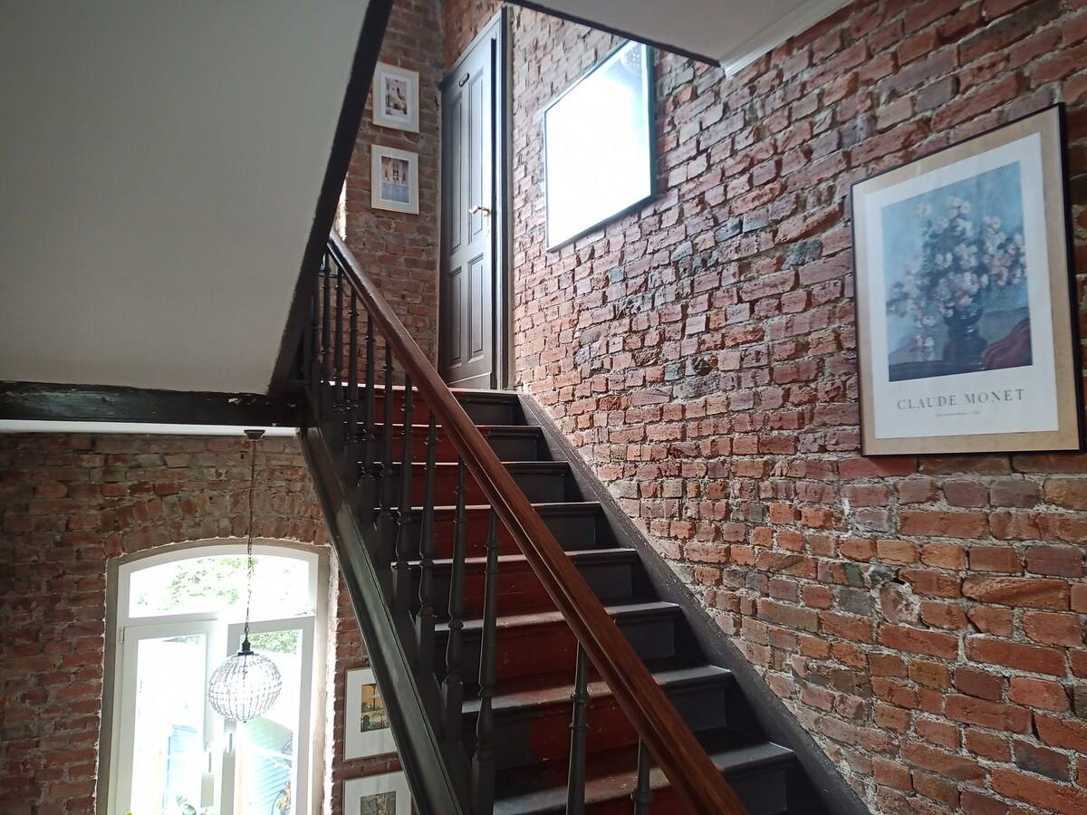 A staircase with a dark wooden railing leads to an upper floor, framed by exposed brick walls. Artworks, including a Claude Monet print, are displayed on the wall. A door at the top of the stairs provides access to additional rooms, while large windows allow natural light to fill the space.