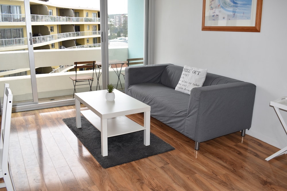 A cozy living area features a grey sofa and a white coffee table, placed on light wooden flooring. A small plant decorates the table. Large glass sliding doors lead to a balcony, allowing natural light to fill the space.