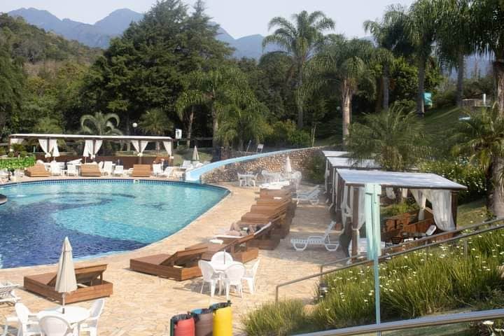 A large outdoor pool is surrounded by palm trees and lounge chairs. Relaxation areas with shaded cabanas are visible alongside the pool. Scenic mountains serve as a backdrop, contributing to a serene atmosphere.