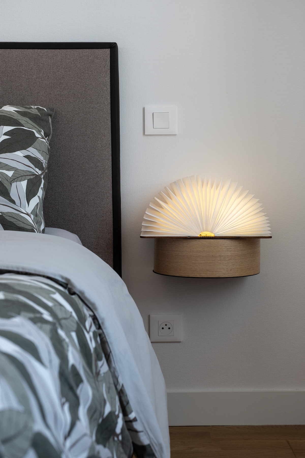 A bedside lamp with a unique fan-shaped design emits a soft glow, mounted beside a bed with patterned pillows. The neutral tones of the wall and bedding create a calm environment, while the wooden base adds a touch of warmth.
