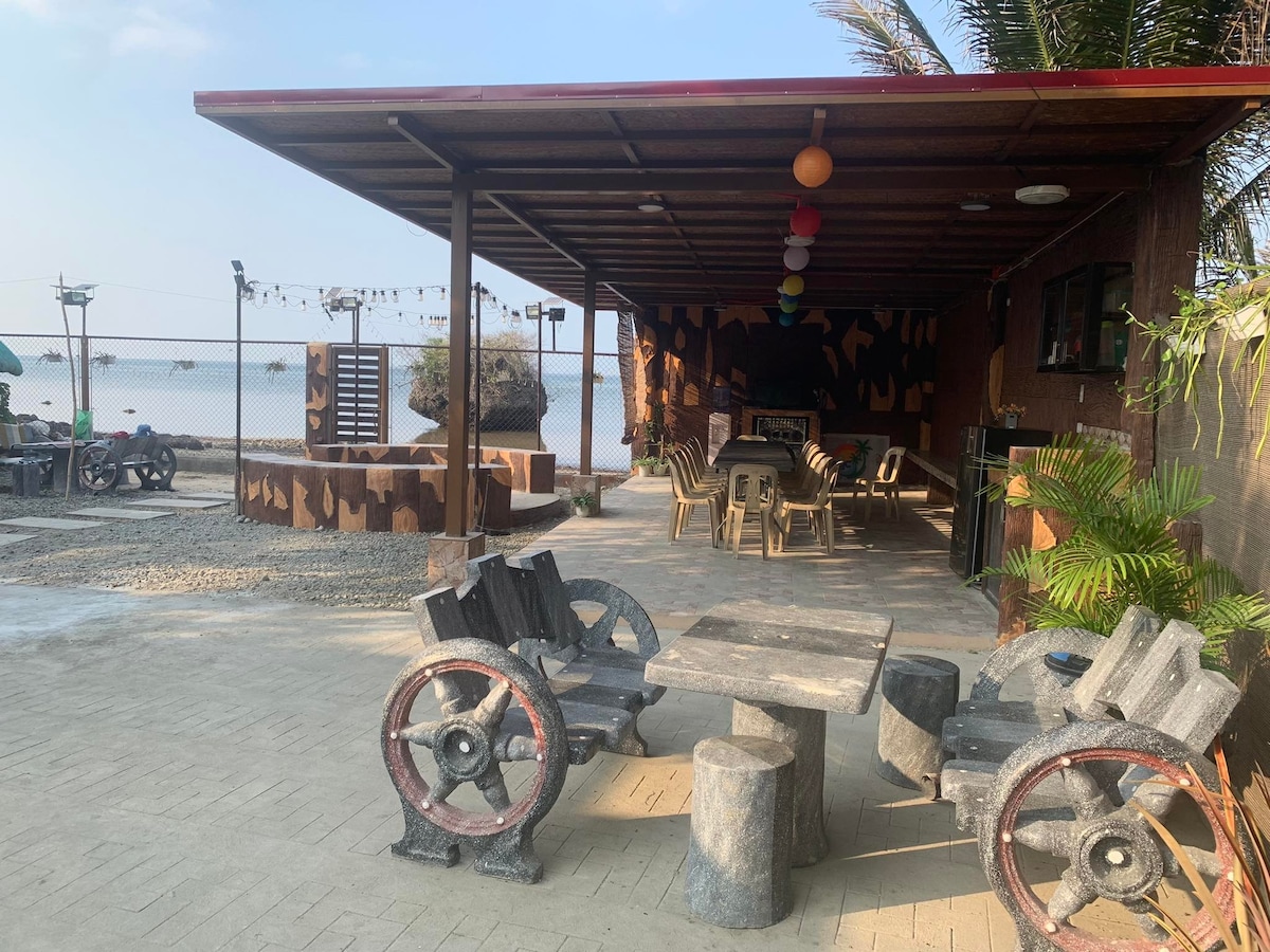 An outdoor seating area is highlighted, featuring a large table surrounded by unique chairs resembling decorative wheels. A shaded space is visible with a backdrop of the beach, complemented by string lights and colorful decorations hanging from the ceiling.