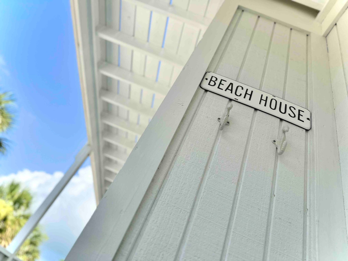 A sign labeled 'BEACH HOUSE' is displayed prominently on a white wooden wall, suggesting an inviting entrance. Clear blue skies are visible above, with hints of palm trees in the background, emphasizing the coastal setting.