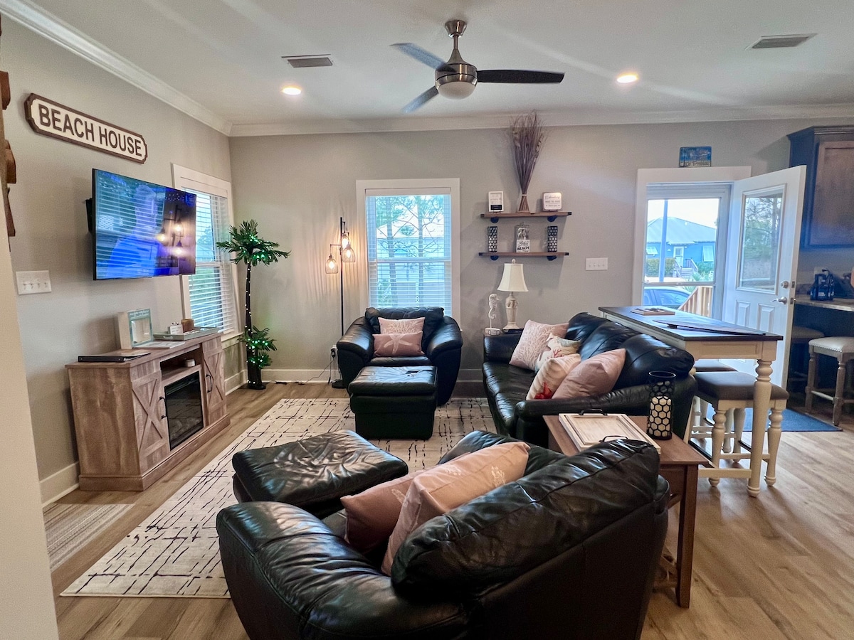 A comfortable living area showcases two dark leather sofas adorned with soft cushions, accompanied by a matching ottoman. A ceiling fan hangs above, while natural light filters through the windows, enhancing the relaxed atmosphere. A television is mounted on the wall, and décor elements are thoughtfully arranged.