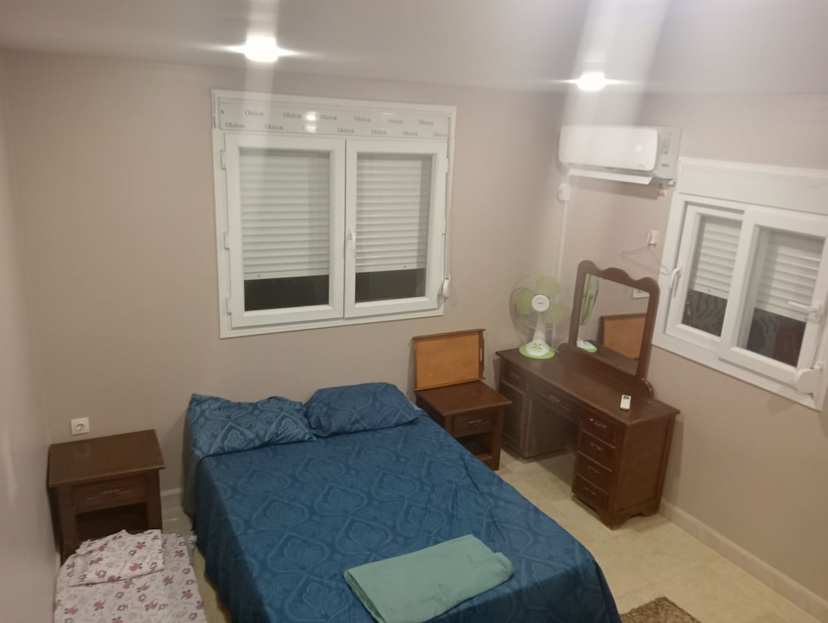 A cozy bedroom features a spacious bed with a blue cover set against neutral walls. Natural light enters through two windows, with striped blinds partially closed. Two wooden nightstands are positioned beside the bed, along with a vintage vanity table and a fan.