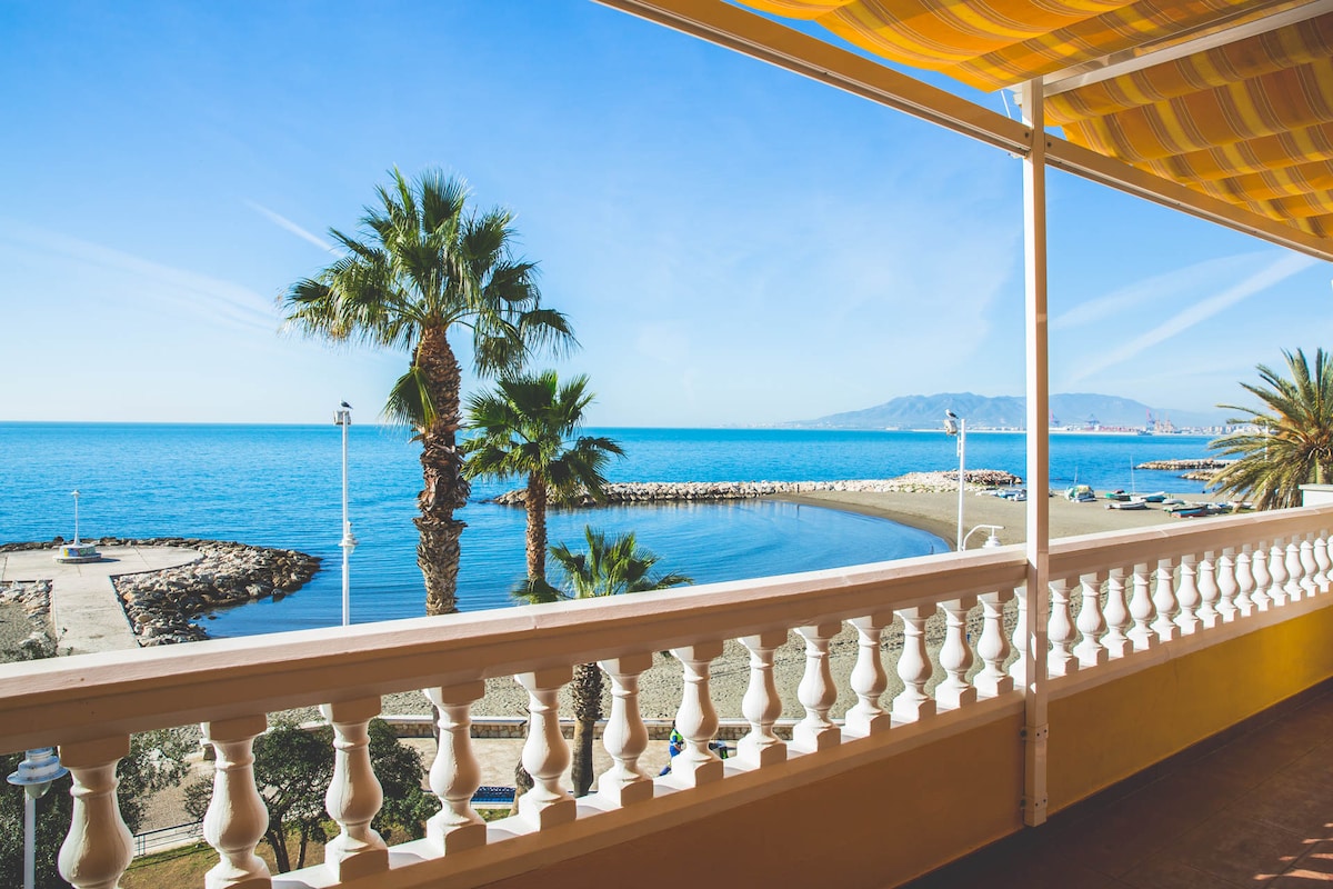 A panoramic view of the ocean is visible from the balcony, framed by vibrant palm trees. The sunlight bathes the water in a bright hue, while the promenade stretches alongside the beach. A clear sky adds to the serene coastal atmosphere.