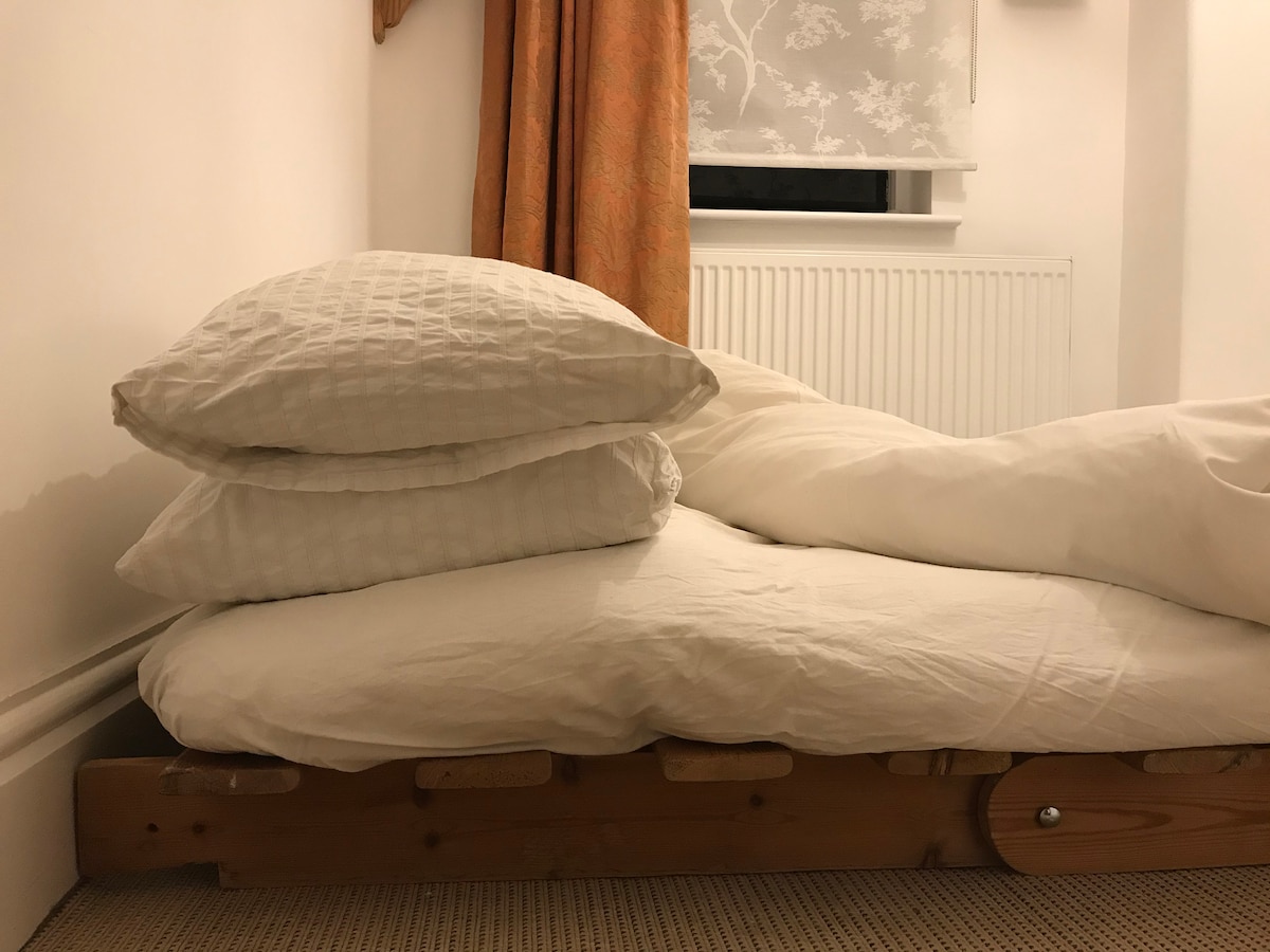 A comfortable futon bed is positioned against a wall, featuring two stacked pillows and a white duvet. Natural light filters through a partially concealed window, while a light-colored curtain adds softness to the space.