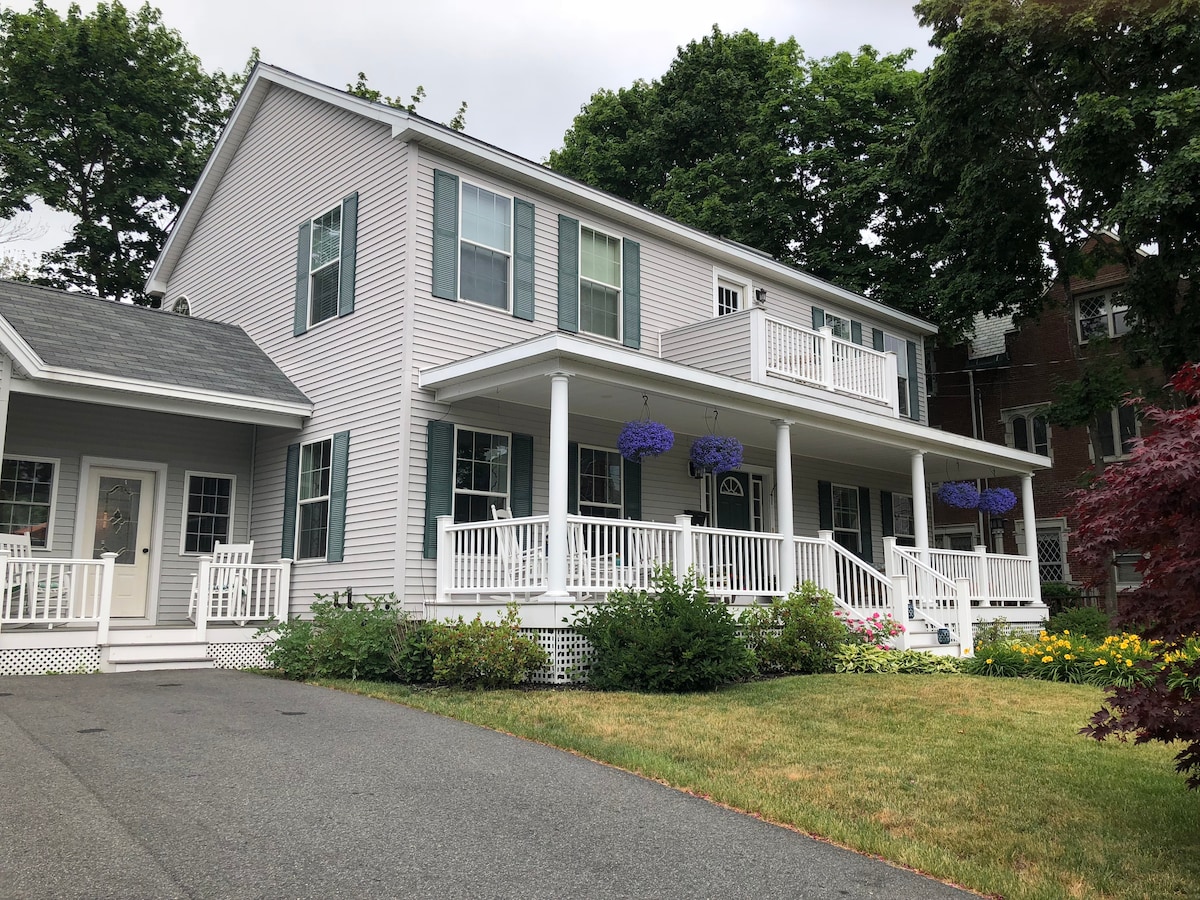 A two-story house is presented with light grey siding and multiple large windows adorned with white trim. A welcoming front porch features a white railing, while purple flowers hang gracefully from it. A well-maintained lawn with colorful flowers adds to the inviting appearance.