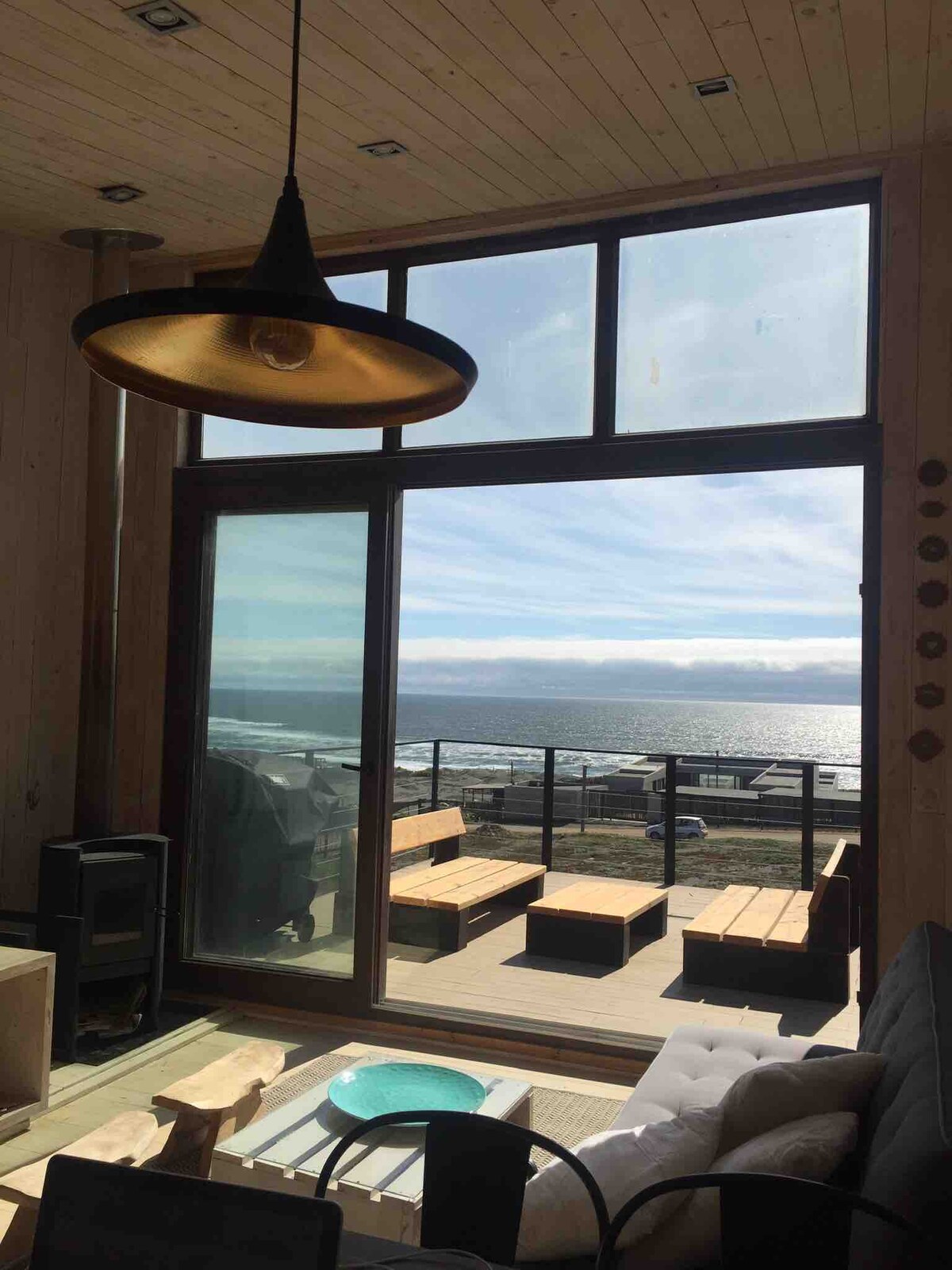 A large glass door opens to a view of the ocean, allowing natural light to fill the room. Comfortable seating is positioned nearby, with wooden benches visible outside on a spacious terrace, creating a seamless connection between indoor and outdoor spaces.
