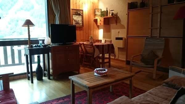 A cozy living area showcases a wooden coffee table, surrounded by light-toned furniture. A flat-screen television sits on a side table, and a comfortable seating arrangement faces a large window, allowing natural light to enter. A decorative rug adds warmth to the wooden floor.