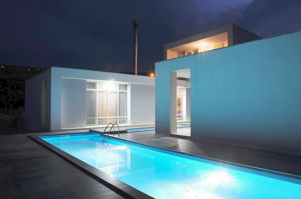 A modern villa is showcased at dusk, with a sparkling pool at the forefront. The pool area is illuminated with soft blue lighting, reflecting against the sleek white exterior of the building. Large glass doors provide a glimpse of the interior living space.