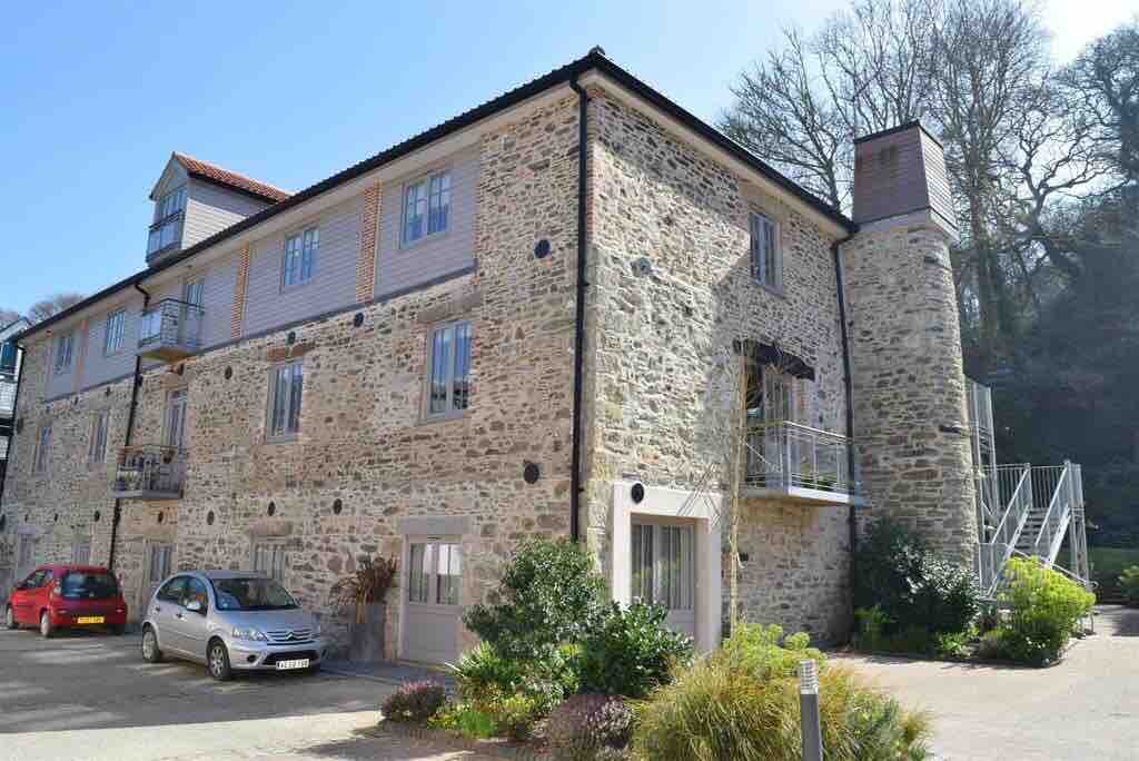 The exterior of the stone building reflects traditional architecture with a modern touch. Large windows provide natural light, while a small parking area accommodates vehicles. Surrounding greenery enhances the setting, and trees can be seen in the background, contributing to the peaceful atmosphere.