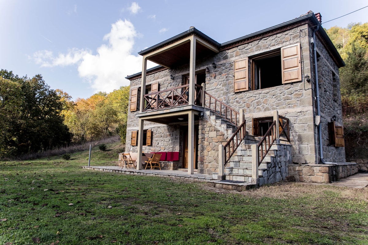 A two-story stone house is set against a natural backdrop, featuring wooden shutters and a wide staircase leading to the upper balcony. Outdoor seating is arranged near the entrance, complemented by lush greenery and trees surrounding the property.