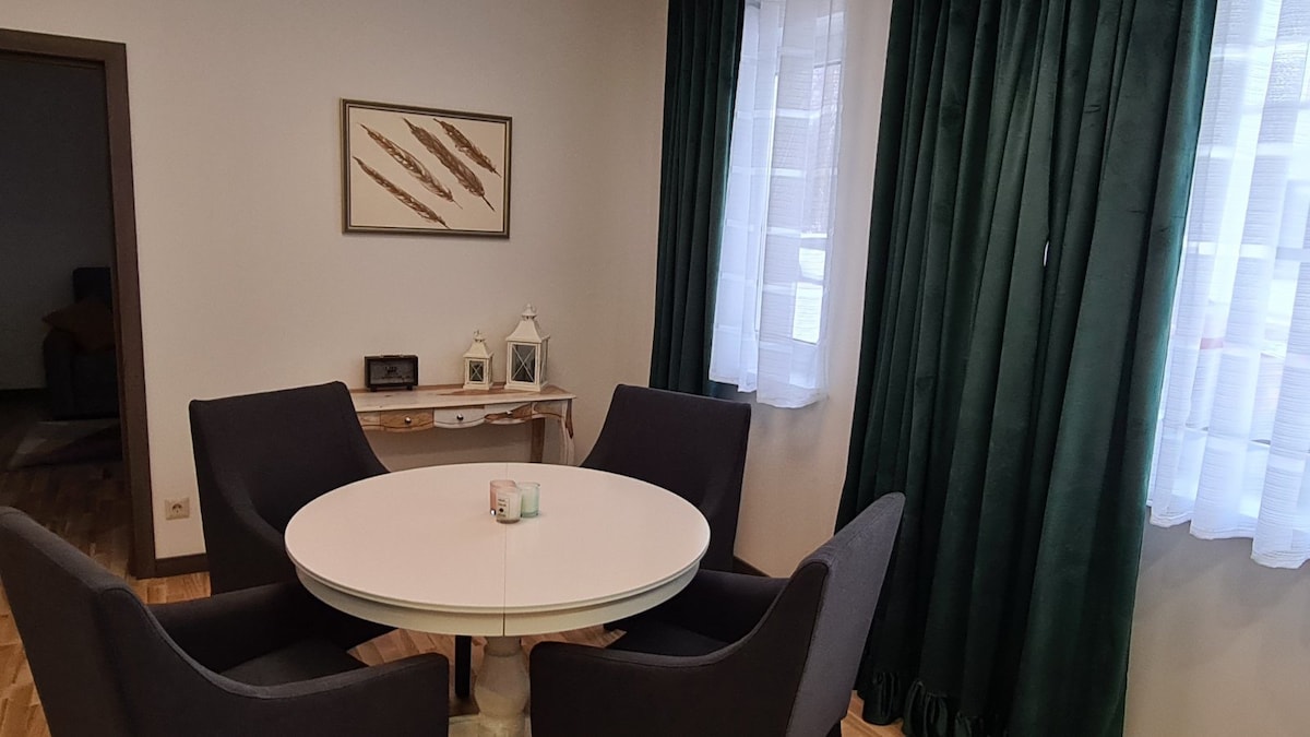 A round dining table is surrounded by four dark upholstered chairs. Green curtains frame the windows, allowing natural light to filter in. A decorative wall piece designed with natural elements hangs above a console table that features lanterns and a small speaker.