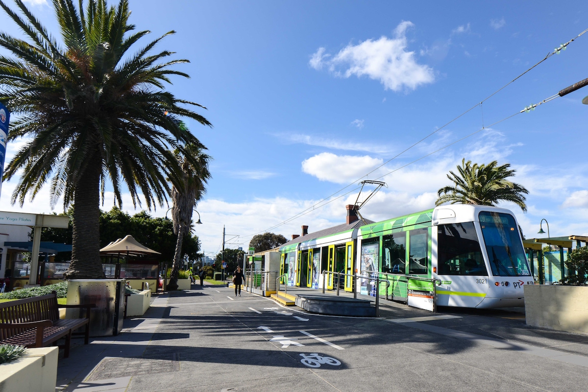 A light rail tram is positioned at a stop along a palm tree-lined promenade. Benches and pathways indicate a welcoming area for pedestrians. The vibrant sky features scattered clouds, providing a bright atmosphere for leisurely strolls near the beach.