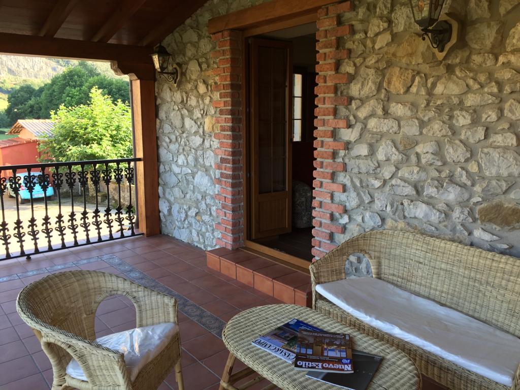 A stone terrace features a seating area with a woven sofa and armchair, complemented by a small table. Lush greenery is visible in the background, and large wooden doors lead into the interior, allowing natural light to brighten the space.